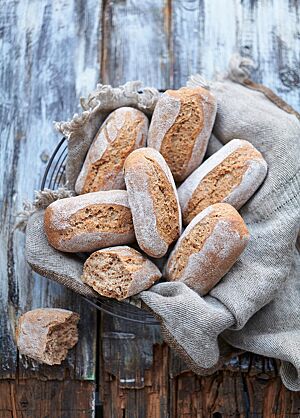 Die Dinkelchen liegen in einem mit einem grob gewebtem grauen Tuch ausgelegtem Drahtkorb. Bei einem auseinandergebrochenen Brötchen wird die kleinporige, elastische Krume sichtbar.