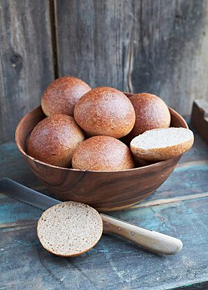 Die glänzenden, goldbraun ausgebackenen Burgerbuns liegen in einer Holzschale. Ein aufgeschnittener Burgerbun lässt die wattige, kleinporige Krume erkennen, die von einer feinen Kruste umgeben ist.