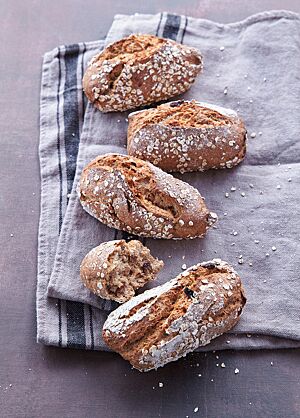 Die mit Haferflocken bestreuten Müslaner liegen auf einem grauen Tuch. Ein auseinandergebrochenes Brötchen zeigt die elastische, saftige, kleinporige Krume.
