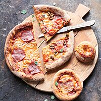 Pizzen und Pizzasnacks in verschiedenen Größen und mit unterschiedlichen Belägen liegen zum Teil geschnitten auf einem runden Holzbrett.