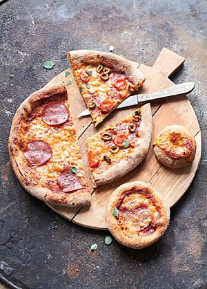 Pizzen und Pizzasnacks in verschiedenen Größen und mit unterschiedlichen Belägen liegen zum Teil geschnitten auf einem runden Holzbrett.