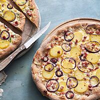 Zwei knusprig gebackene, mit Kartoffeln, Zwiebeln, Speck und Schmand belegten Schnäcks, liegen auf runden Holzbrettern. Neben einem geschnittenen Schnäck liegt ein Wellenschliffmesser mit Holzgriff.