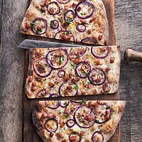 Der gedrittelte, mit Zwiebeln, Speck und Schmand belegte Vlammkuchen liegt auf einem rustikalen Holzbrett.