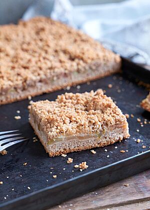 Der angeschnittene Blechkuchen liegt auf einem schwarzen Backblech. Im Anschnitt ist eine Schichtung des feinporigen Bodens, der saftigen Apfelschicht und der goldbraunen Streusel deutlich erkennbar.