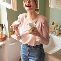 Eine Studentin hält glücklich eine mit Butter bestrichene Scheibe Vollkornnussbrot und eine Tasse in der Hand.