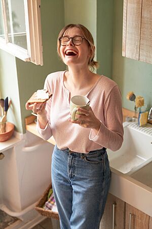 Eine Studentin hält glücklich eine mit Butter bestrichene Scheibe Vollkornnussbrot und eine Tasse in der Hand.