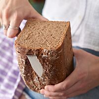 Von einem im Kasten gebackenen Dreikornbrot mit ganzen Getreidekörnern in der Krume wird mit einem Messer eine Scheibe abgeschnitten.