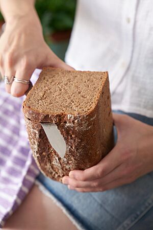Von einem im Kasten gebackenen Dreikornbrot mit ganzen Getreidekörnern in der Krume wird mit einem Messer eine Scheibe abgeschnitten.