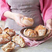 Goldgelb ausgebackene, rustikal aufgerissene Weizenbrötchen liegen in einer Schale. Daneben liegt ein auseinandergebrochenes Brötchen und zeigt die weiche, lockere Krume.