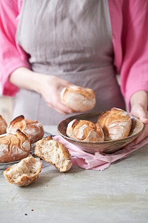 Goldgelb ausgebackene, rustikal aufgerissene Weizenbrötchen liegen in einer Schale. Daneben liegt ein auseinandergebrochenes Brötchen und zeigt die weiche, lockere Krume.