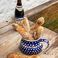 In einem blau-weiß gemusterten Krug stehen vier kräftig ausgebackene Haager Bierstangen neben einer Flasche Weißbier.