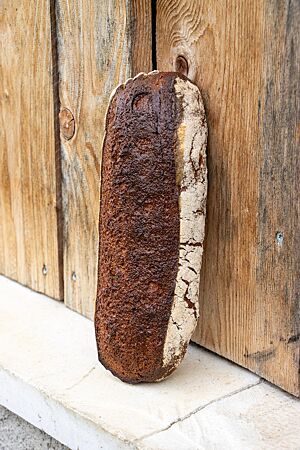 Ein schmales Buchebener Heidebrot lehnt kräftig ausgebacken aufrecht an einer Holzwand. Seitlich ist die Kruste stark bemehlt, auf der Oberfläche dagegen leicht glänzend und dunkelbraun ausgebacken.
