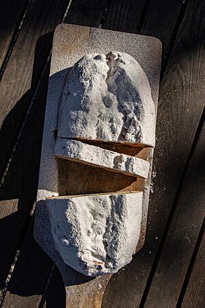Ein reichlich mit Puderzucker bestreuter Stollen liegt angeschnitten auf einem Holzschieber in der Sonne.