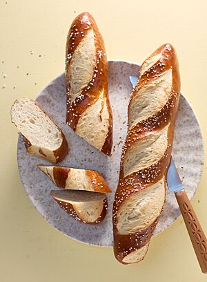 Zwei mit Brezelsalz bzw. Sesam bestreute Laugenstangen in der Größe von Baguettes liegen auf einem Teller. Die angeschnittene Laugenstange XL (links) zeigt die kleinporige, fast weiße Krume.