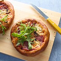 Zwei kleine Laugenpizzen liegen auf einem Holzbrett. Sie sind mit Tomatensauce, Käse, Salami, Pilzen und frischem Rucola belegt.